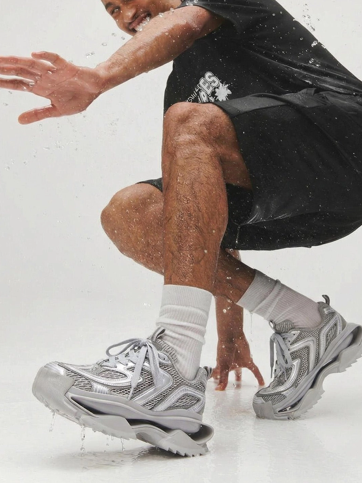 Silver mesh running sneakers worn with black sportswear in a studio water splash scene, highlighting the textured upper and technical runner silhouette