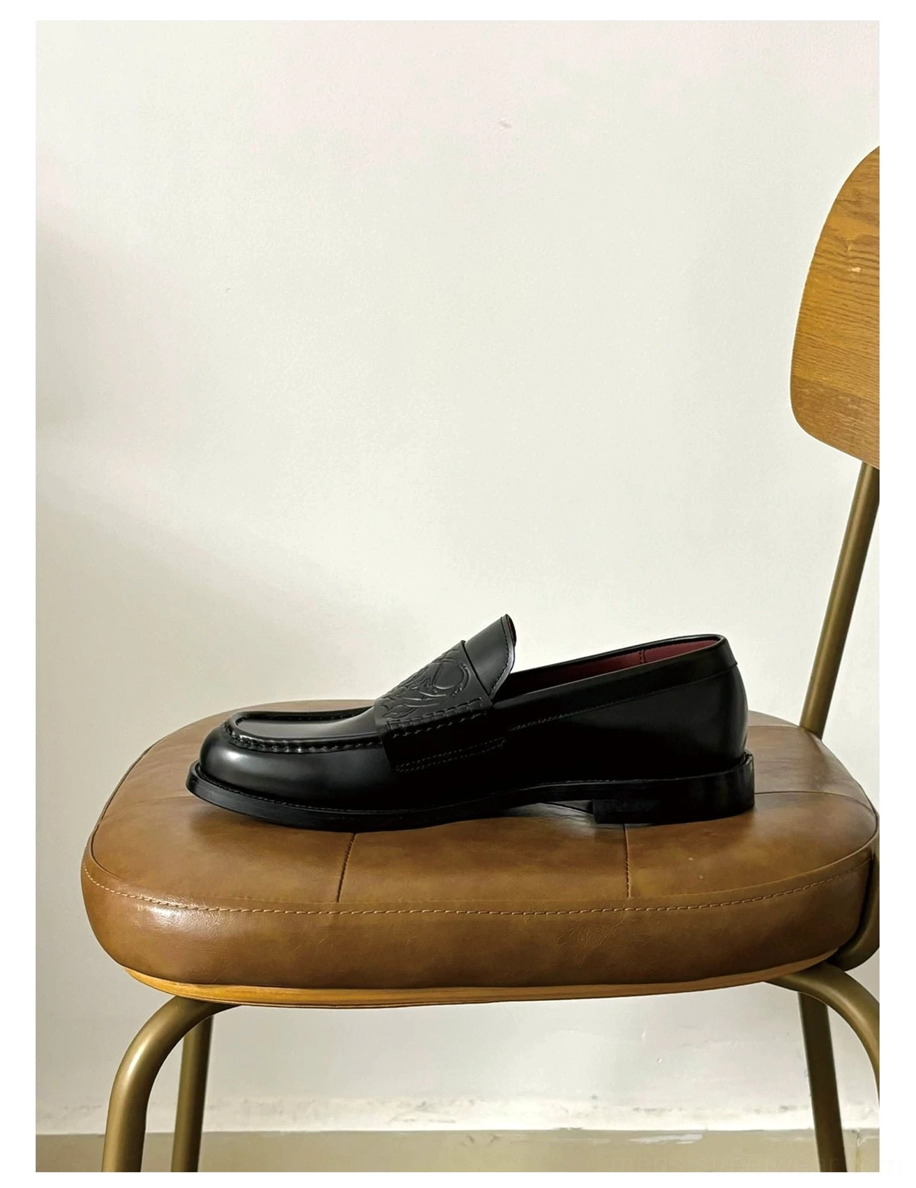 Single black embossed leather loafer displayed on a vintage wooden chair showing its square toe, low heel, and burgundy interior