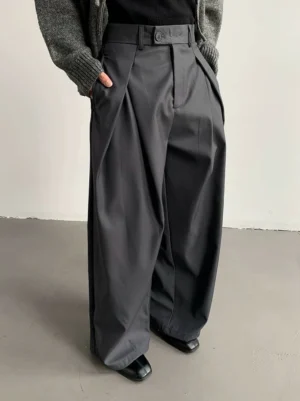 Smoke gray pleated wide-leg trousers close-up showing buttoned waistband front pleats functional pockets and relaxed tailored silhouette