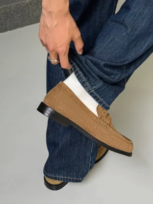 Tan khaki square-toe suede penny loafers worn with dark denim and white socks, showing the black rubber outsole and low rear side profile
