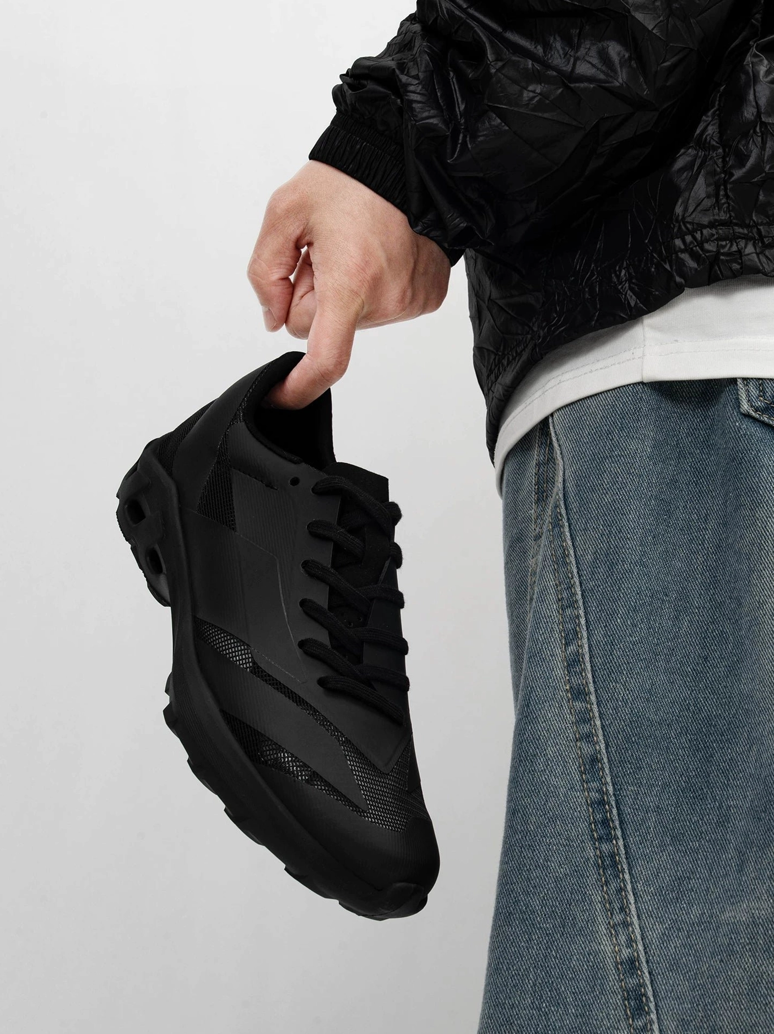 Streamlined black techwear sneakers held against a casual outfit with black jacket and blue jeans, showing lace-up closure and textured matte upper