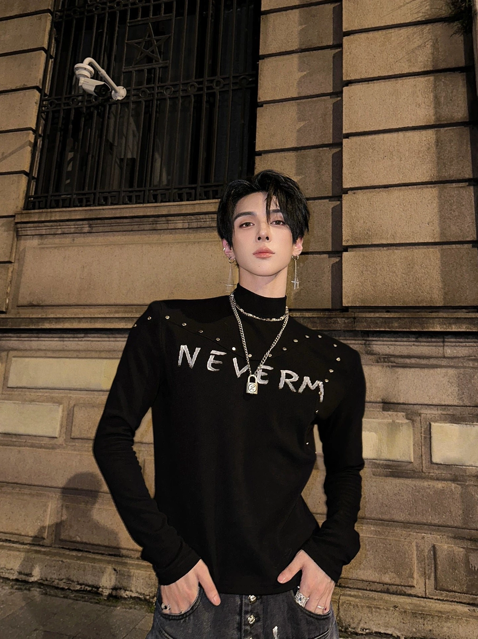 Male model wearing a black studded gothic knit turtleneck sweater with distressed chest graphic, slim long sleeves, printed jeans, and lock pendant necklace on a retro stone street