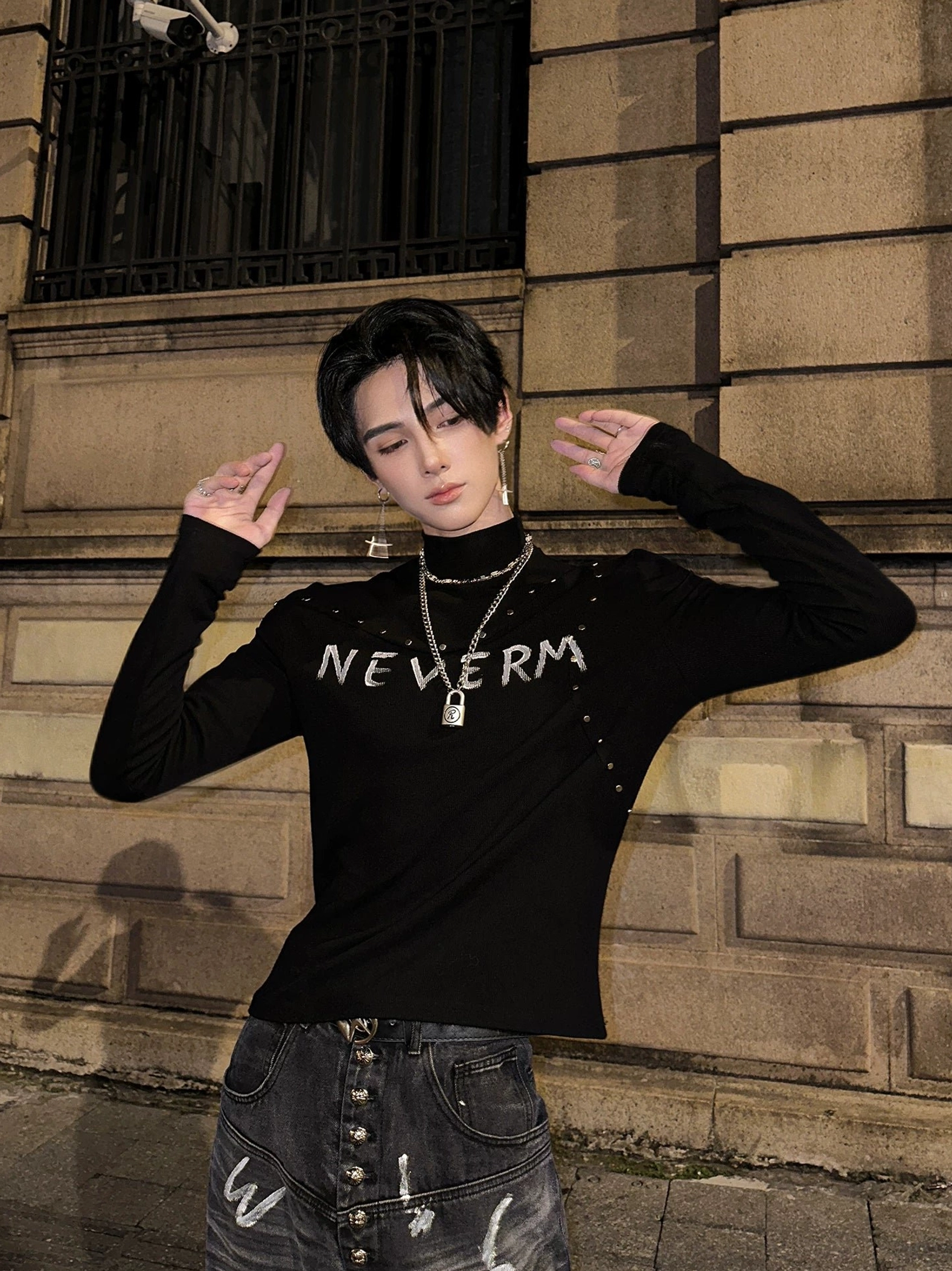 Dynamic front portrait of a male model wearing a studded gothic turtleneck knit sweater with graphic print, slim sleeves, printed jeans, and pendant necklace