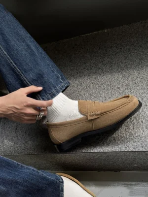 Tan suede square-toe penny loafer shown from an overhead side angle with dark jeans, white socks, visible stitching, and silver rings