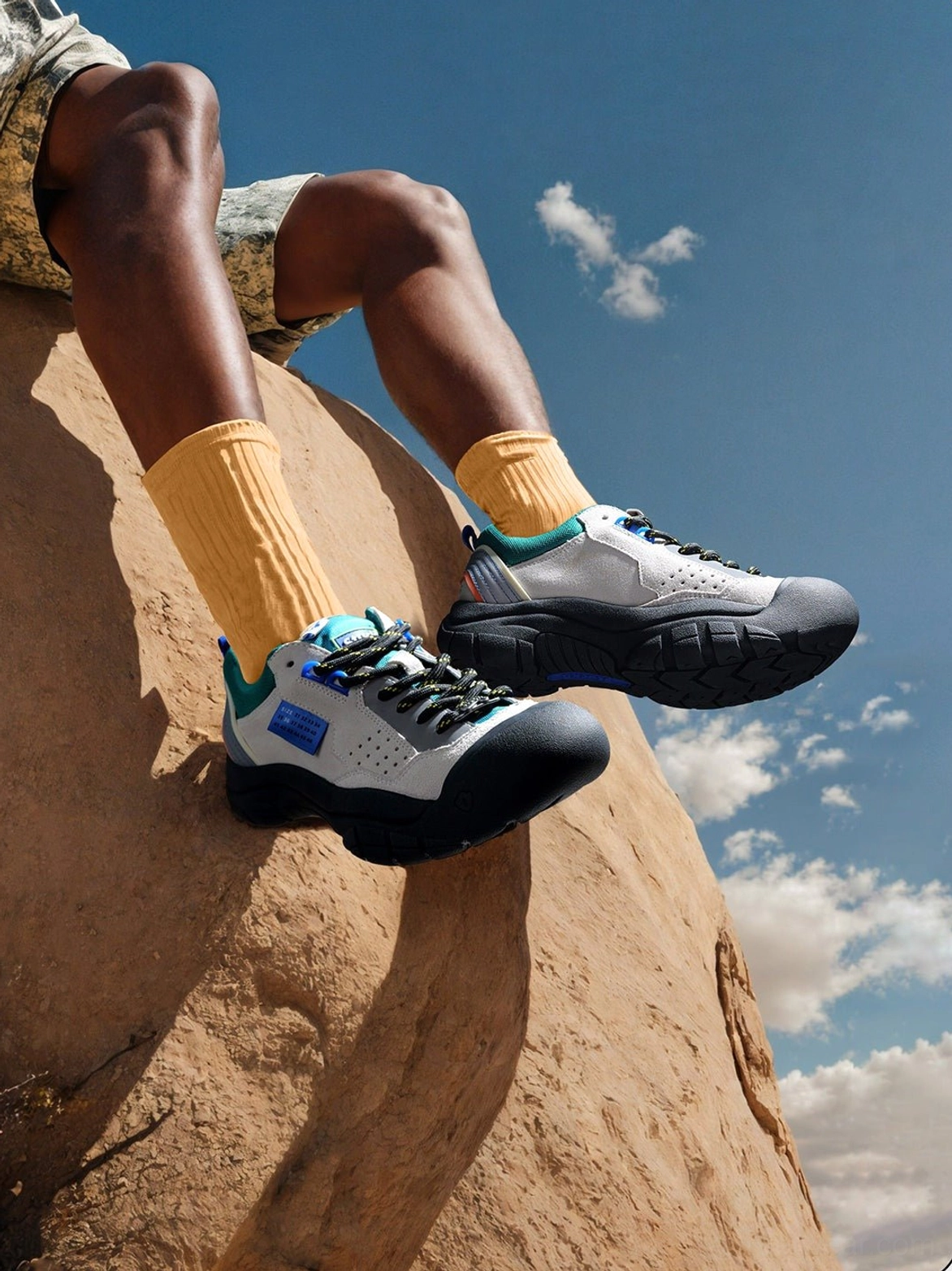Male streetwear model wearing off-white teal suede texture trail platform sneakers with yellow socks and camo shorts on rugged desert terrain