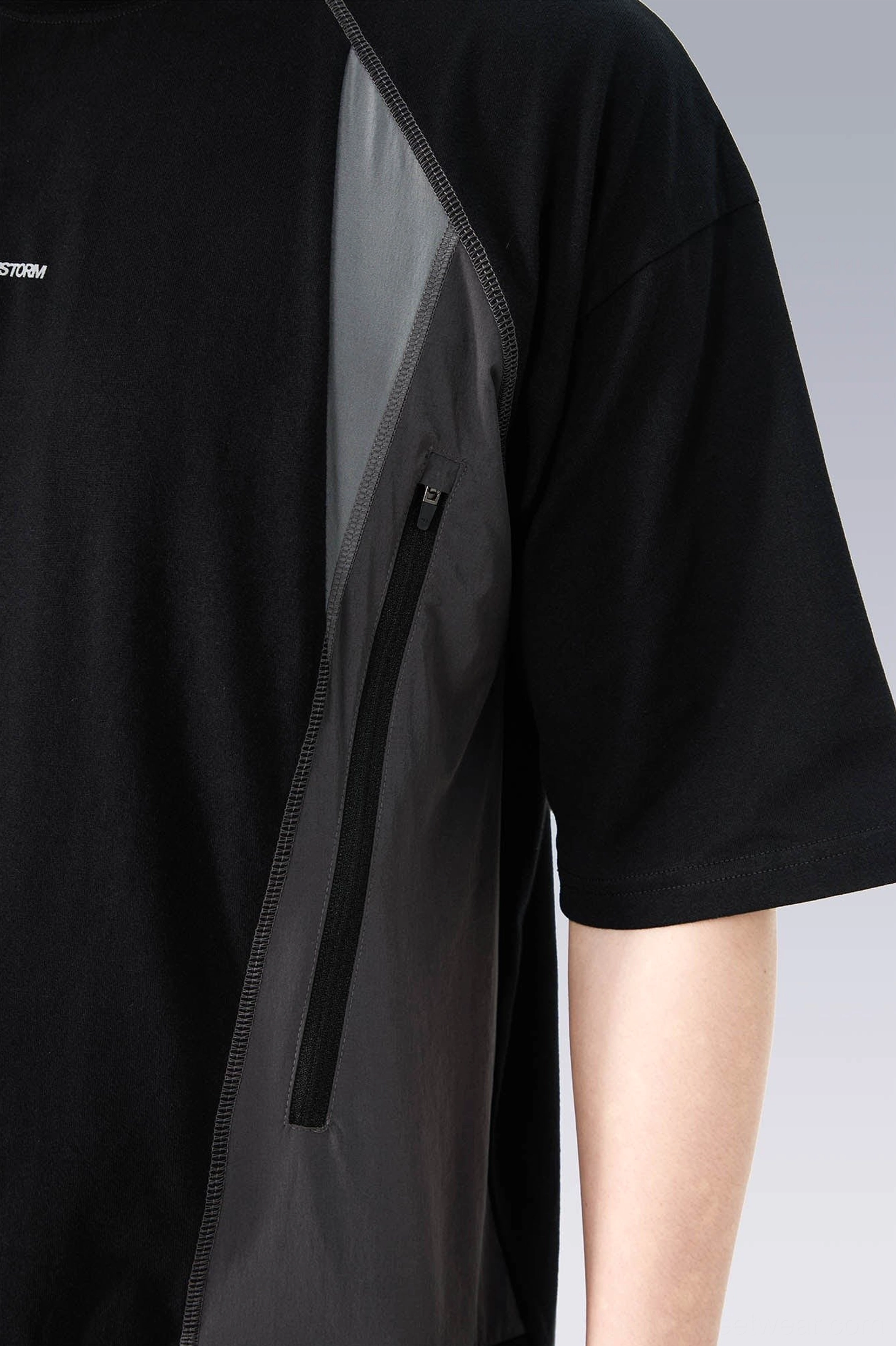 Close-up of the technical side panel on a black streetwear T-shirt with contrasting grey fabric, vertical zipper pocket, and tonal stitching