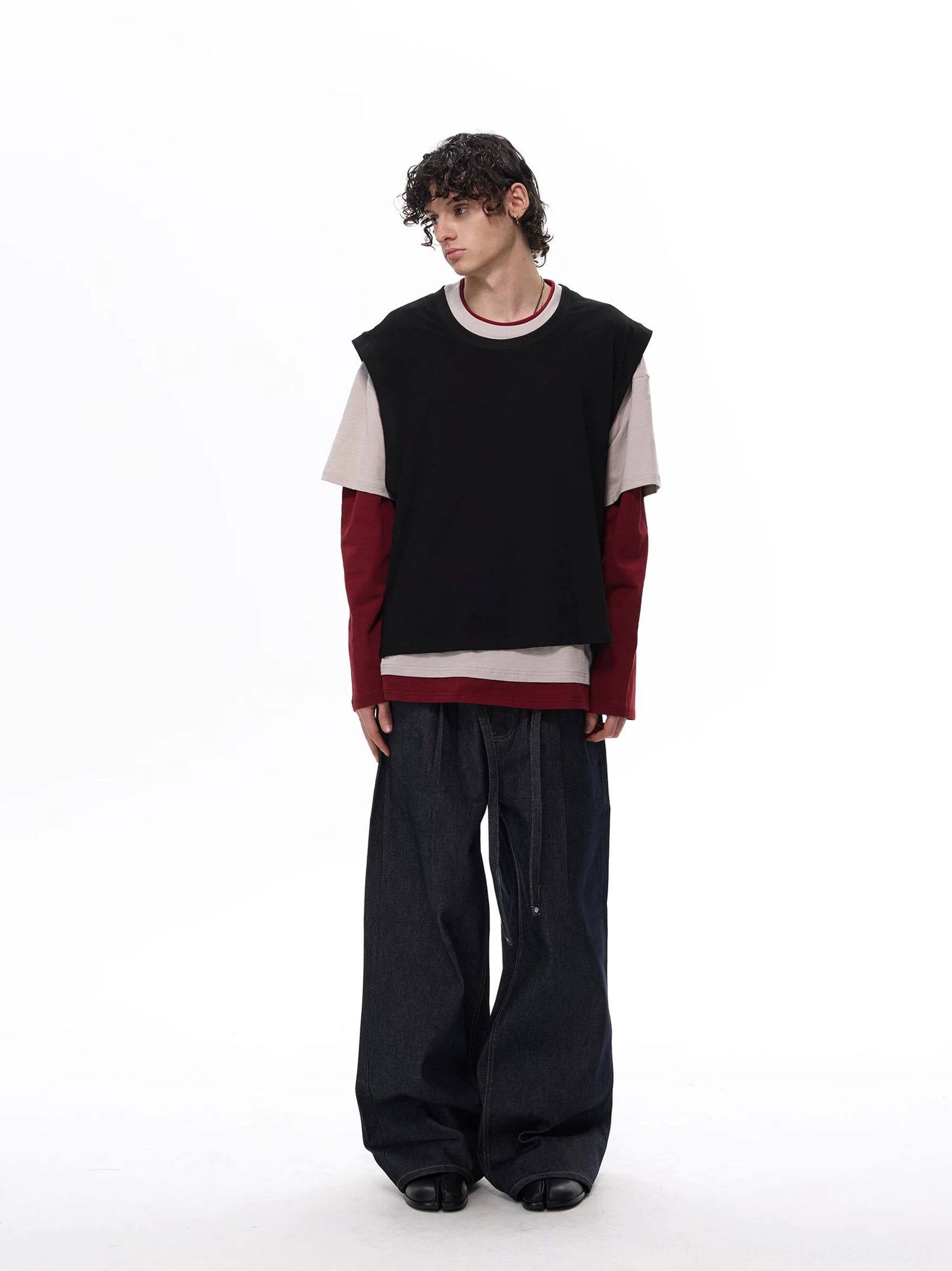 Rear view of a male model wearing a black beige and burgundy two-tone layered vest tee top with wide-leg dark denim in a minimalist streetwear studio