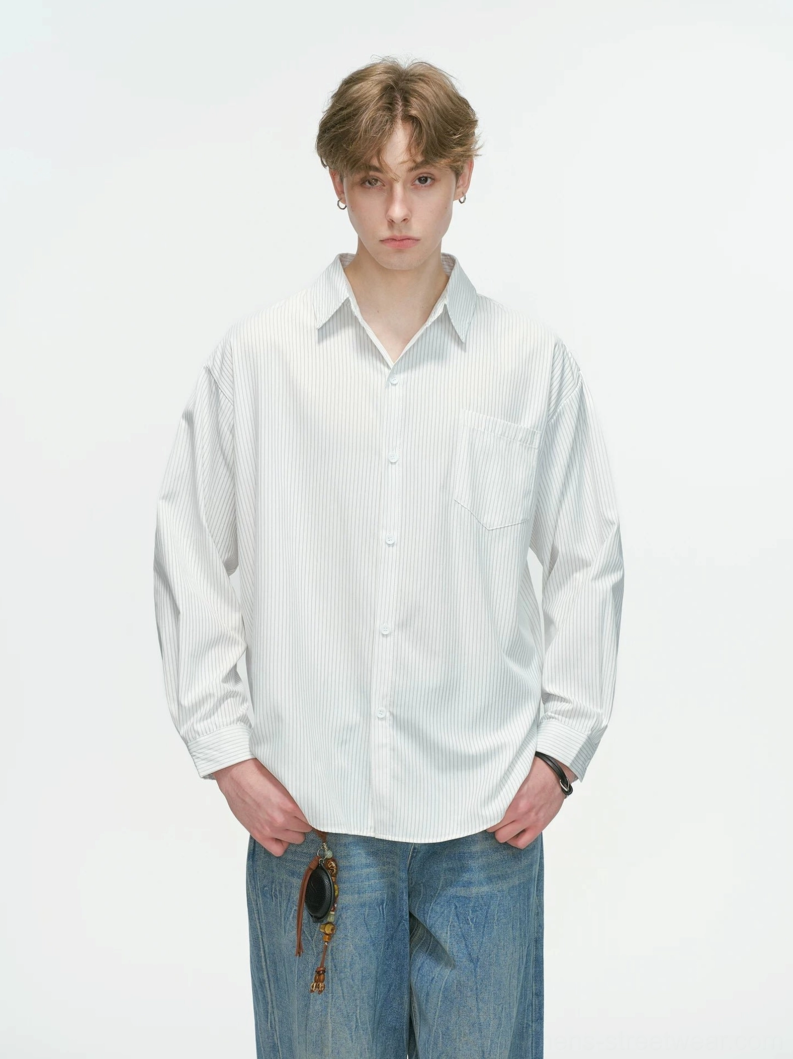 Male model wearing a white vertical stripe relaxed oversized overshirt with chest pocket, wide-leg blue jeans, and sunglasses in a clean studio setting