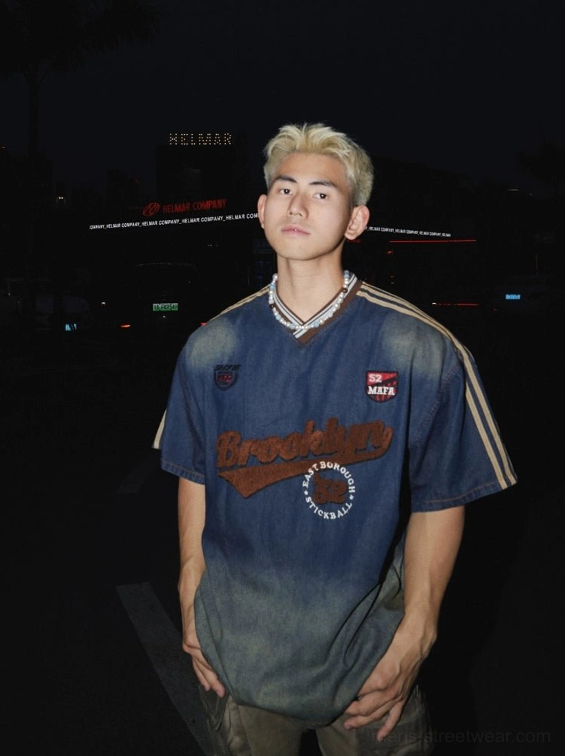 Front view of a vintage blue ombre denim Brooklyn varsity T-shirt with embroidered lettering, athletic patches, striped short sleeves, and contrasting V-neck on an urban streetwear model