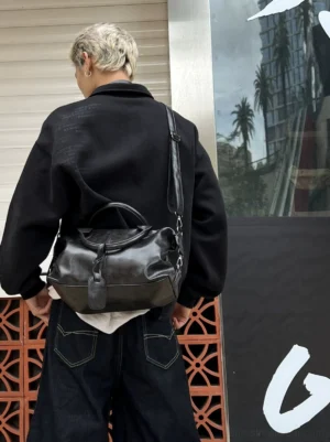 Black vintage faux leather bowling bag worn crossbody with an all-black men's streetwear outfit from the rear