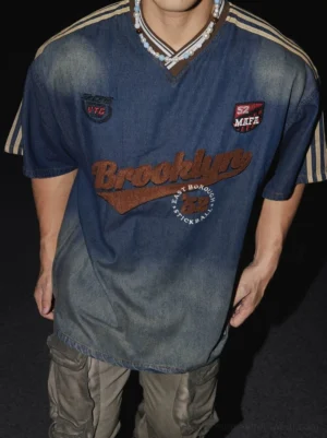 Close-up of relaxed vintage ombre denim-effect T-shirt showing brown ribbed V-neck, embroidered varsity patches, khaki sleeve stripes, and layered streetwear styling