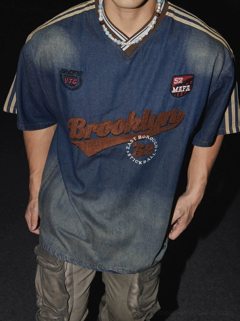 Close-up of relaxed vintage ombre denim-effect T-shirt showing brown ribbed V-neck, embroidered varsity patches, khaki sleeve stripes, and layered streetwear styling