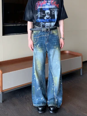 Full front view of faded blue paint splatter wide-leg jeans showing the asymmetric waistband, diagonal seams, and relaxed streetwear fit