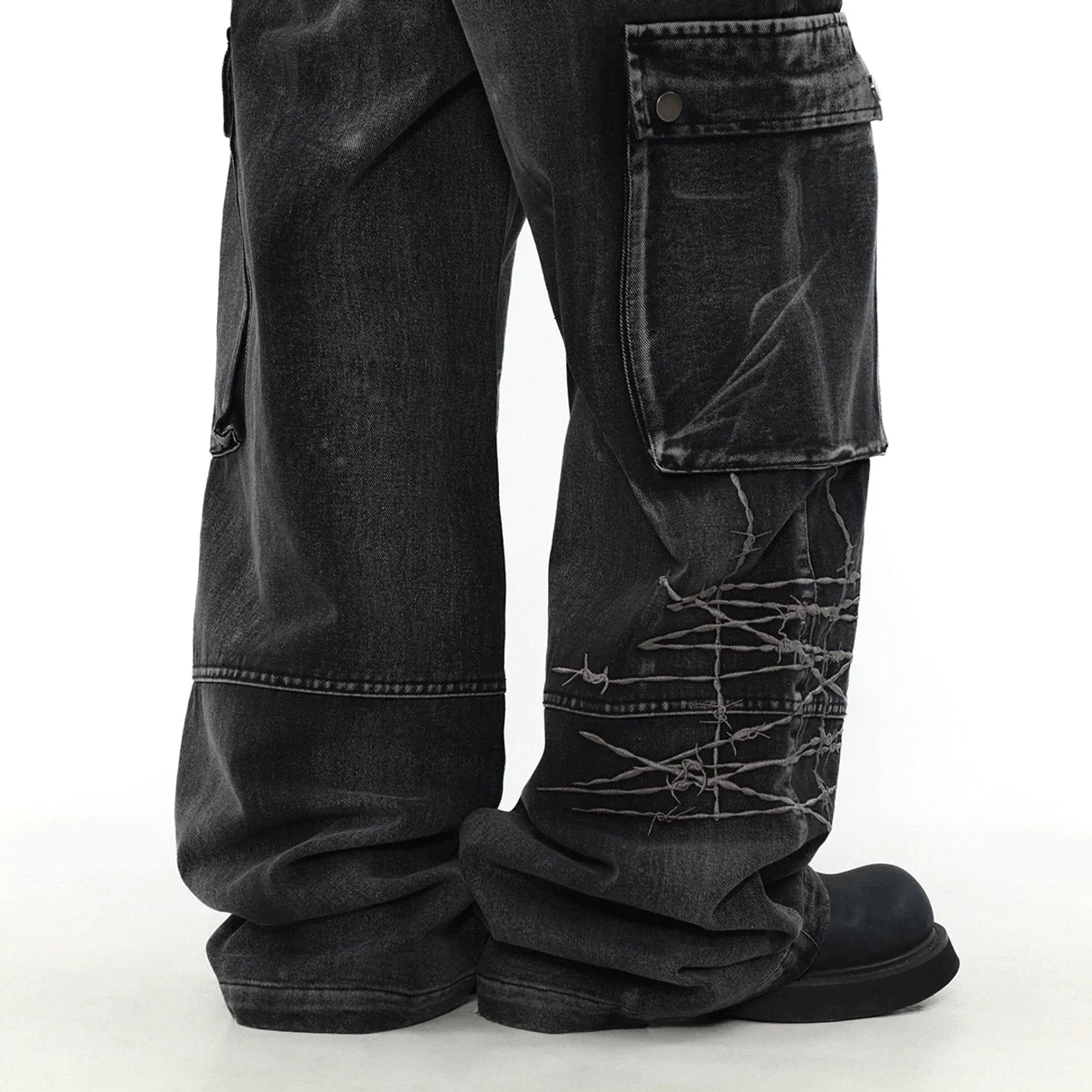 Rear lower detail of washed black wide-leg cargo denim jeans with button-flap pockets, barbed wire stitched graphics, and stacked hems over boots