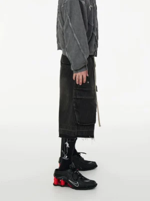 Left side view of faded black washed denim cargo long shorts with exposed side cargo pockets, extended waist drawcords, raw hem, and loose Seoul streetwear fit