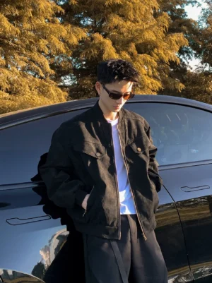 Male model wearing a washed black oversized retro bomber jacket with chest flap pockets beside a black sedan at sunset