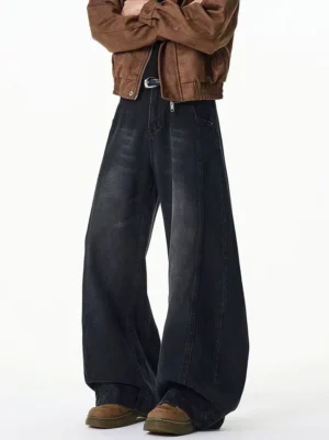 Washed black ultra-wide leg jeans shown from a low-angle front side view with floor-length loose denim legs, metal belt detail, zipper decoration, and brown platform work shoes
