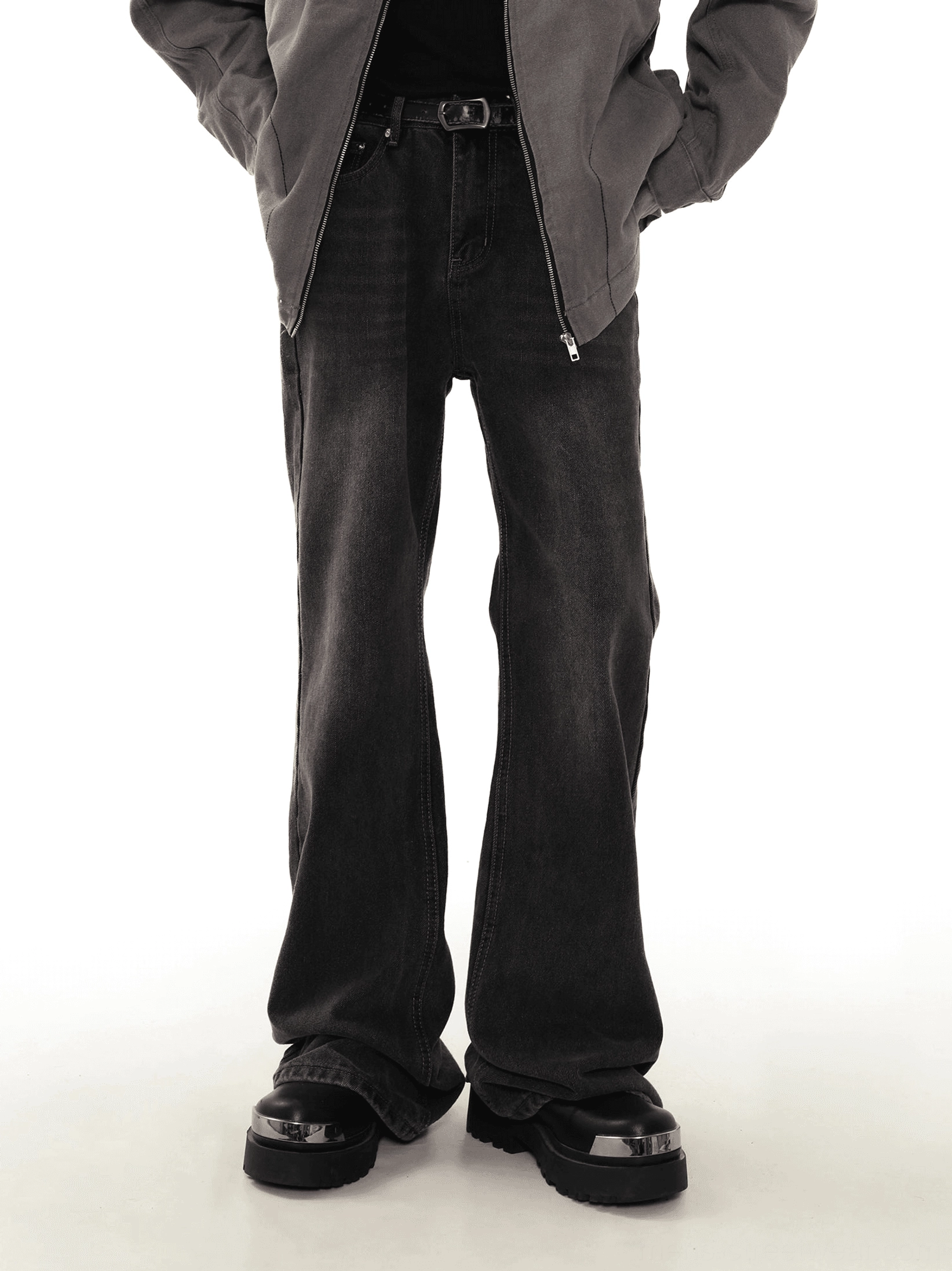 Male model wearing washed black flare jeans with a vintage faded denim finish, gray zip jacket, and platform boots in a white studio