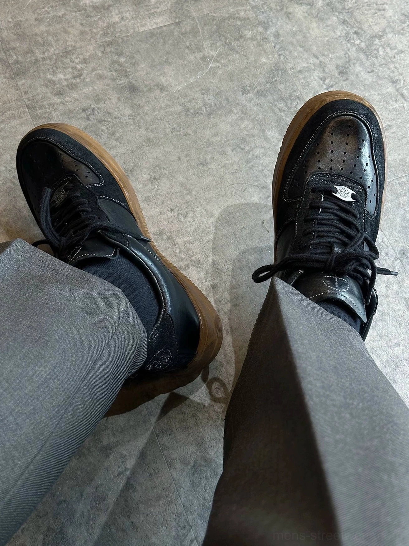 Top view of washed black vintage leather court sneakers worn with grey trousers, featuring metal lace tag, perforated toe box, and brown gum sole