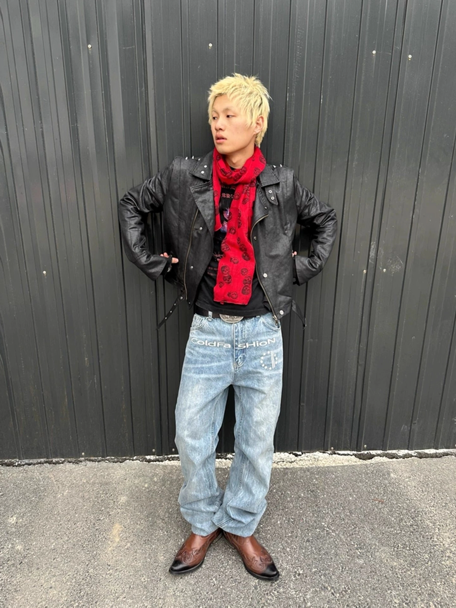 Blonde male model wearing washed blue vintage flare jeans with leather jacket and red scarf against an industrial wall