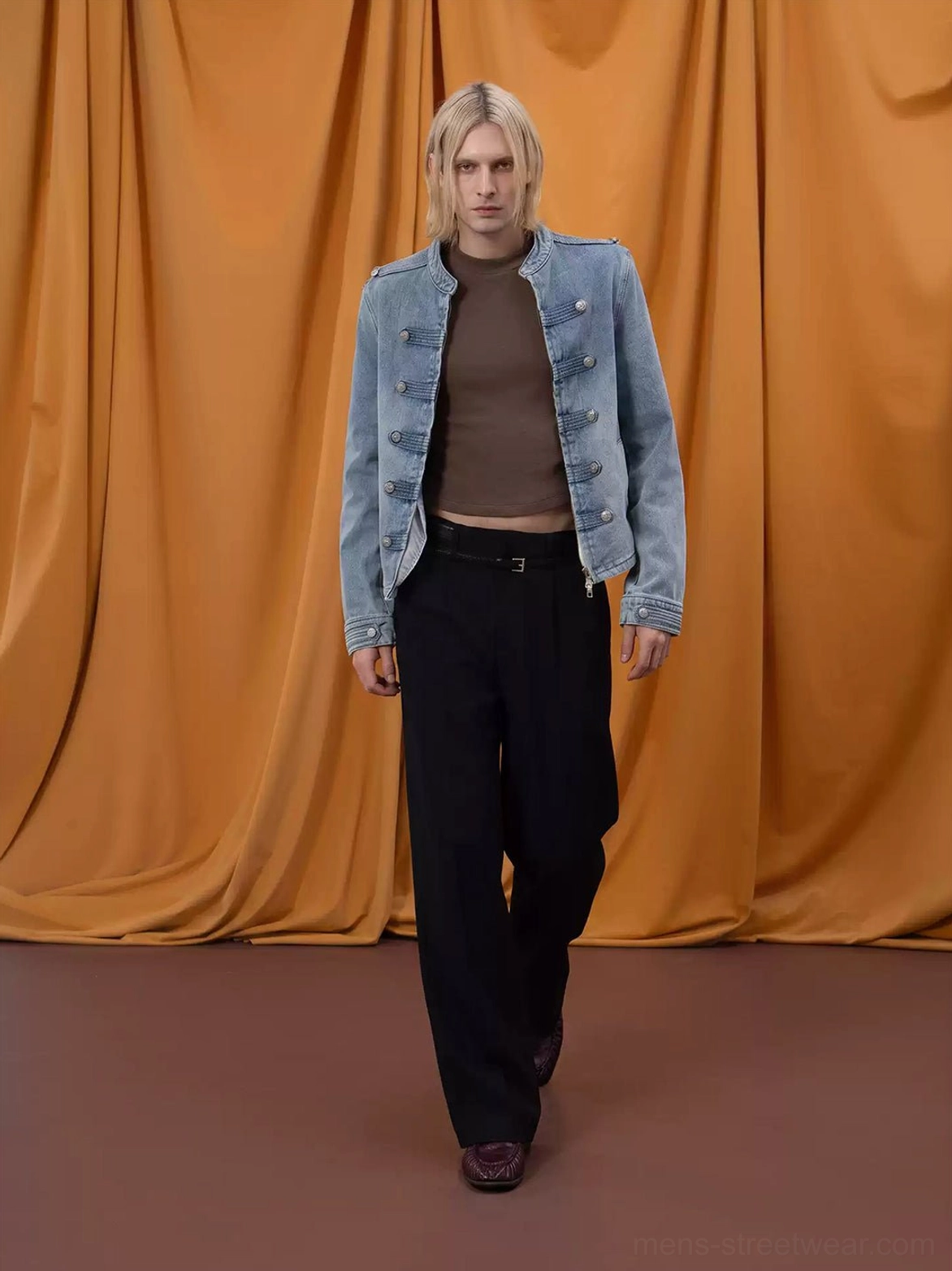 Male model walking in washed denim cropped military zip jacket with silver buttons and black trousers