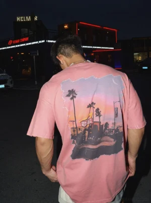 Back view of washed pink oversized streetwear t-shirt with distressed sunset diner and palm tree graphic worn by male model at night