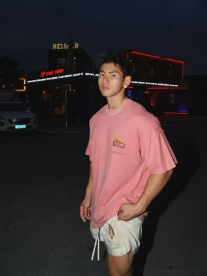 Front view of washed pink oversized crewneck graphic t-shirt with small retro diner chest logo on male model in night street setting