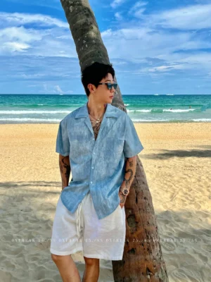 Blue washed seersucker Cuban collar short sleeve streetwear shirt worn with white shorts beside a tropical beach and palm tree