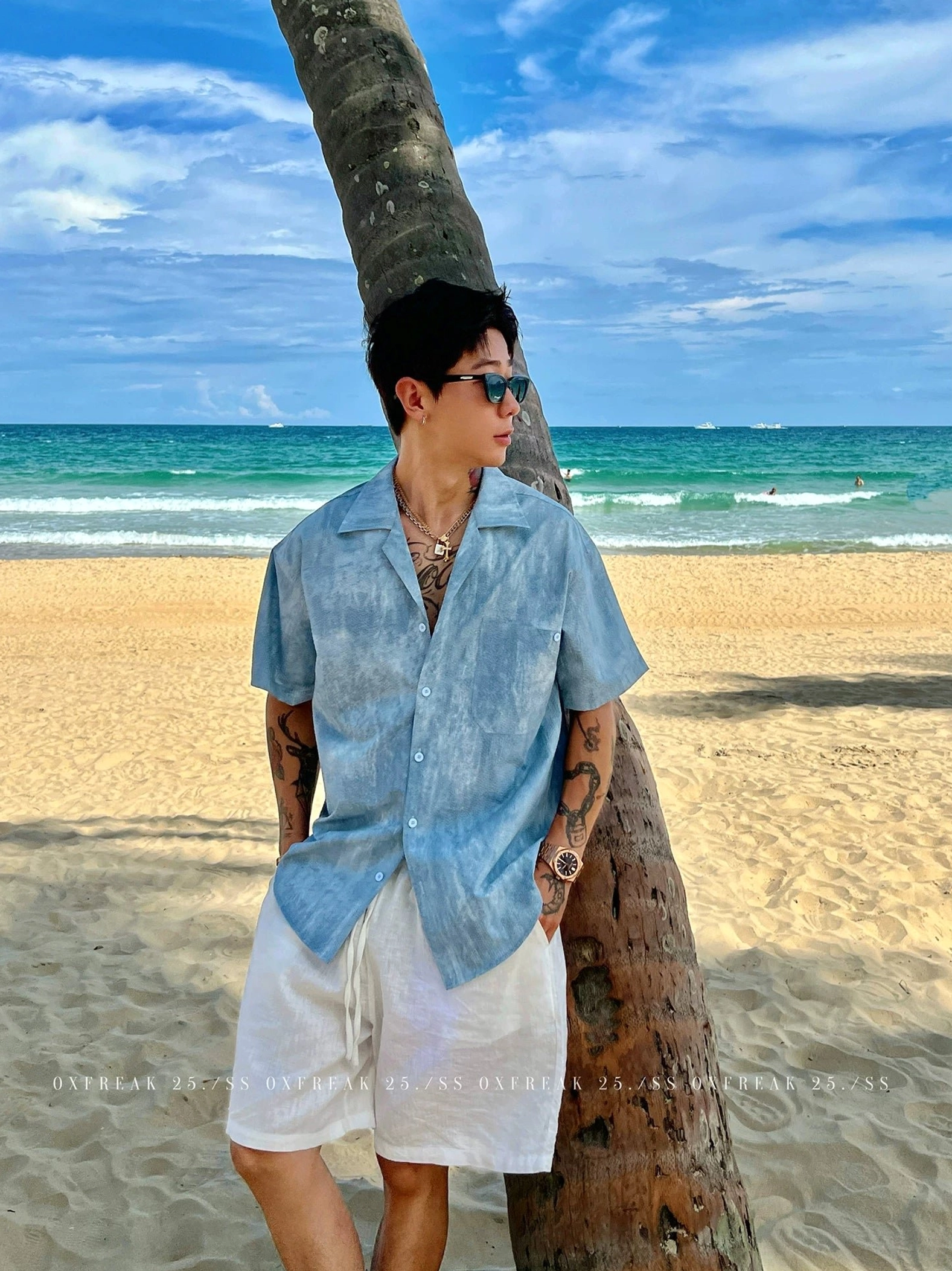 Blue washed seersucker Cuban collar short sleeve streetwear shirt worn with white shorts beside a tropical beach and palm tree