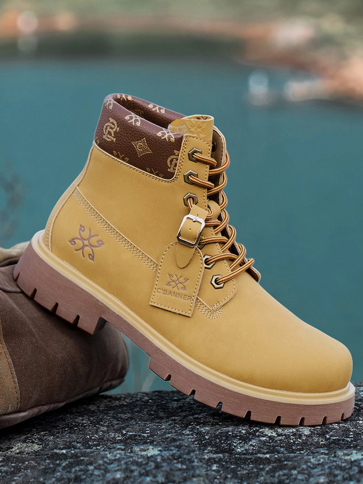 Side three-quarter view of wheat mid-top work boots on seaside rock showing lace-up construction, tongue tag, embossed detail, and thick lug sole