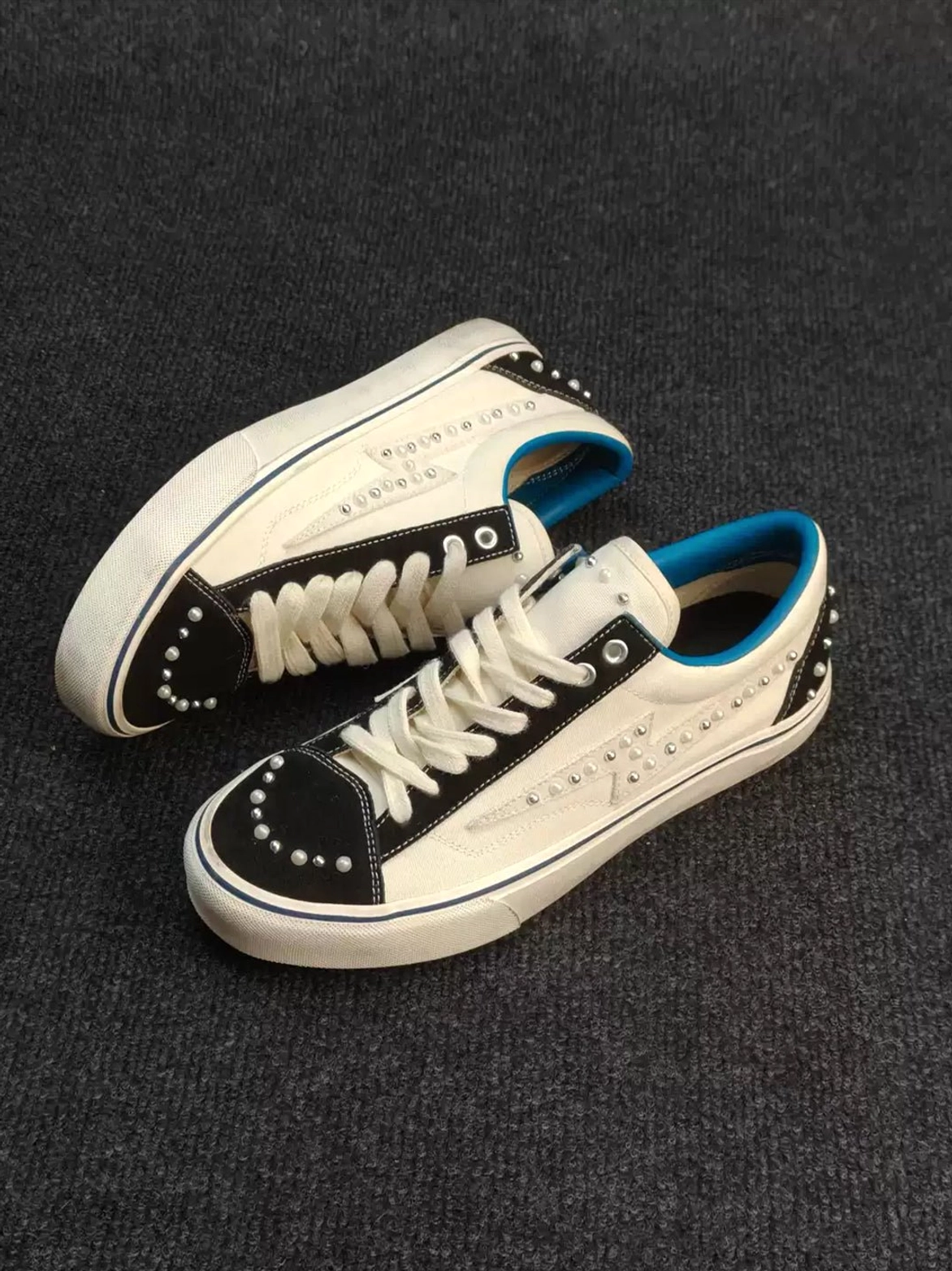 White and black pearl-studded skate sneakers shown from a low oblique angle with blue inner lining, blue midsole trim, and embellished toe and side panels