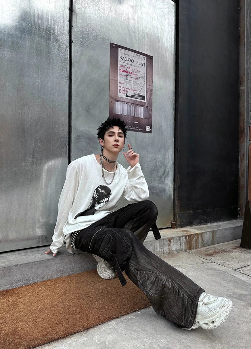 Male model wearing white distressed chunky platform sneakers with wide-leg black pants, a graphic long-sleeve shirt, and silver streetwear accessories