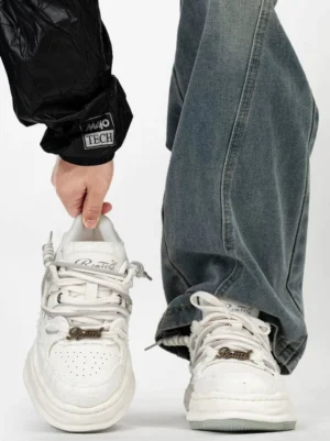 White distressed low-top platform sneakers with layered laces, metal lace tag, chunky melted-edge sole, baggy blue jeans, and black quilted streetwear jacket