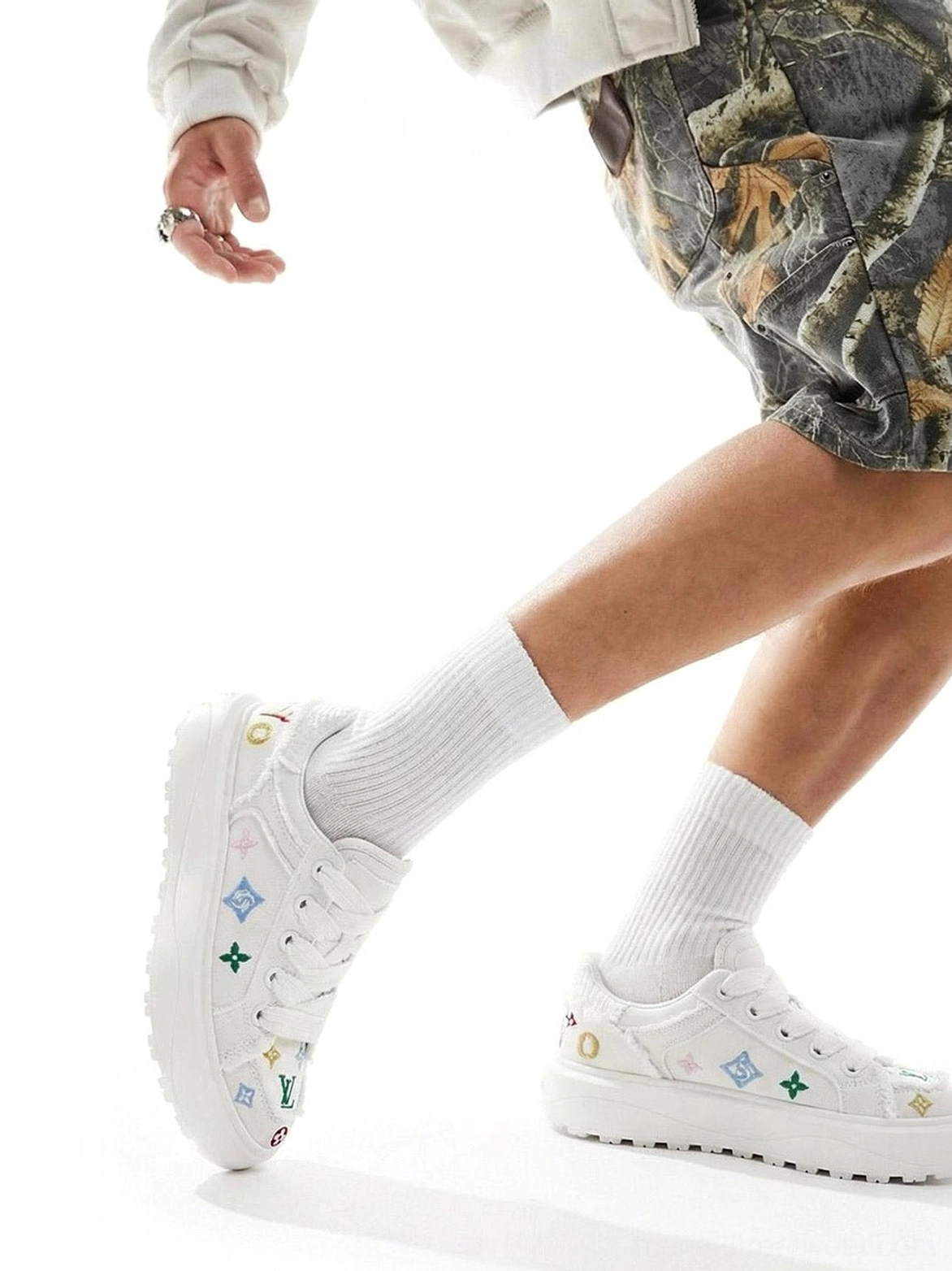 Male model wearing all-white platform skate sneakers with subtle colorful embroidery white crew socks and camouflage shorts in a casual streetwear outfit