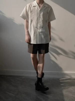 Full body male model in a relaxed white embroidered camp shirt, brown shorts, black socks, and leather boots