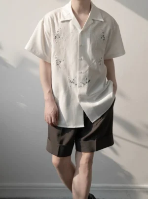 Male model wearing a white floral embroidered camp collar shirt with brown tailored shorts in a sunlit studio