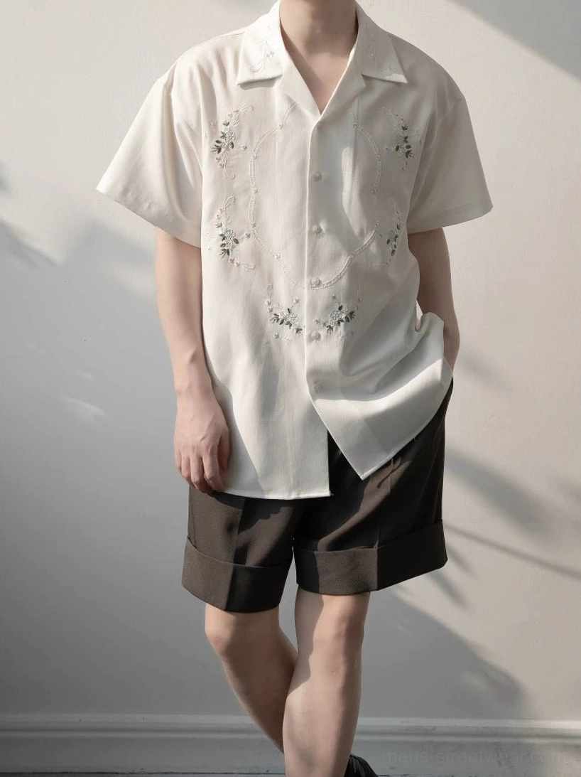 Male model wearing a white floral embroidered camp collar shirt with brown tailored shorts in a sunlit studio