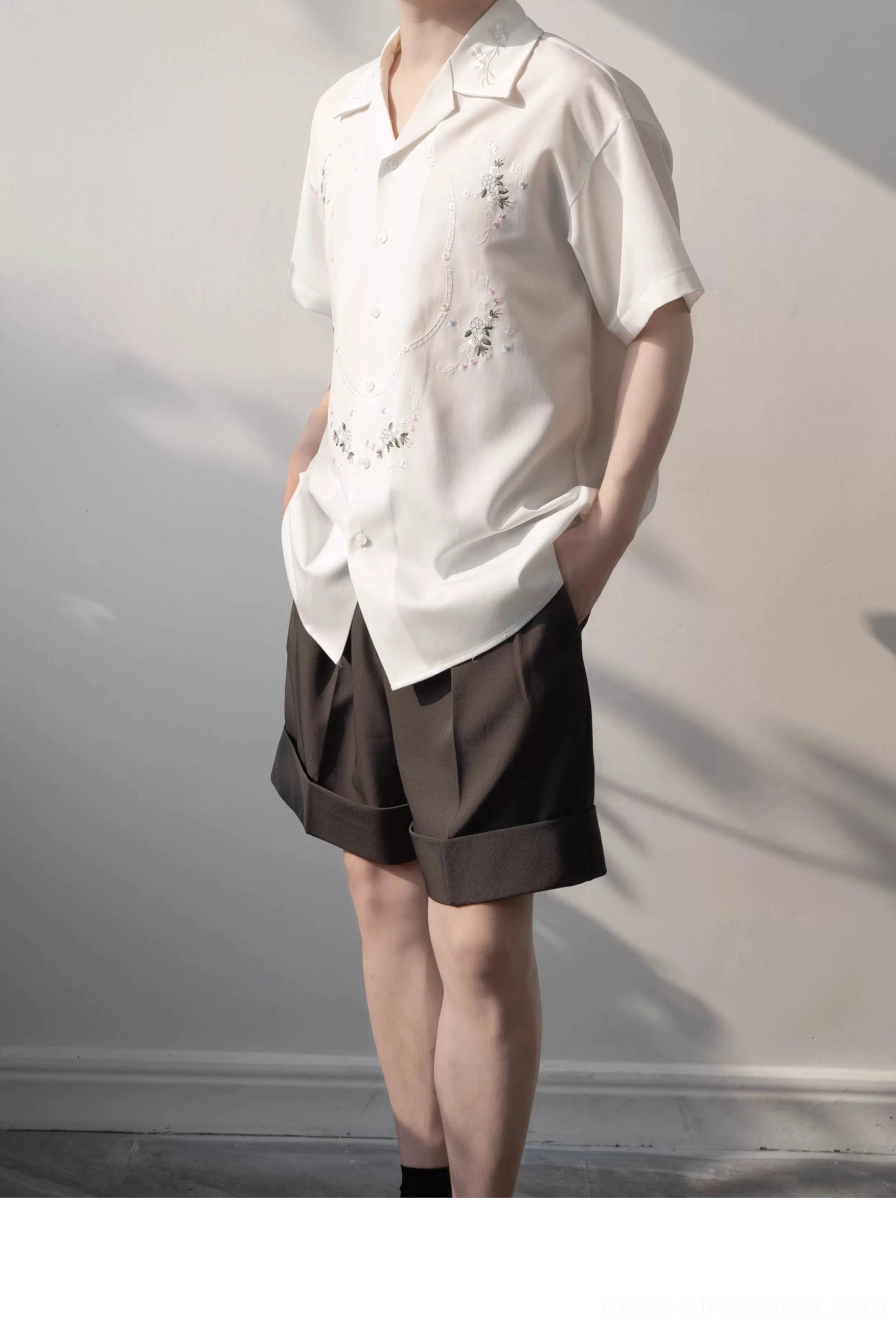 Male model showcasing a relaxed white floral embroidered shirt with high waisted brown tailored shorts