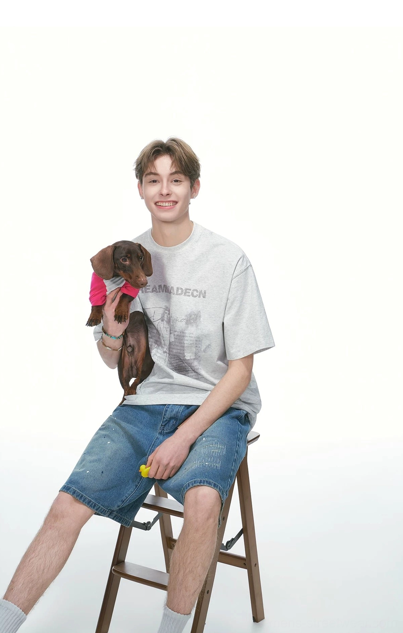 Male model wearing a White Flower Gray oversized graphic streetwear T-shirt with denim shorts while sitting on a wooden stool