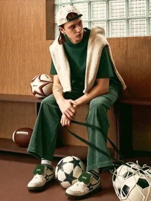 Male model wearing white green detachable tongue streetwear sneakers with a coordinated retro green sport outfit in an indoor athletic lounge