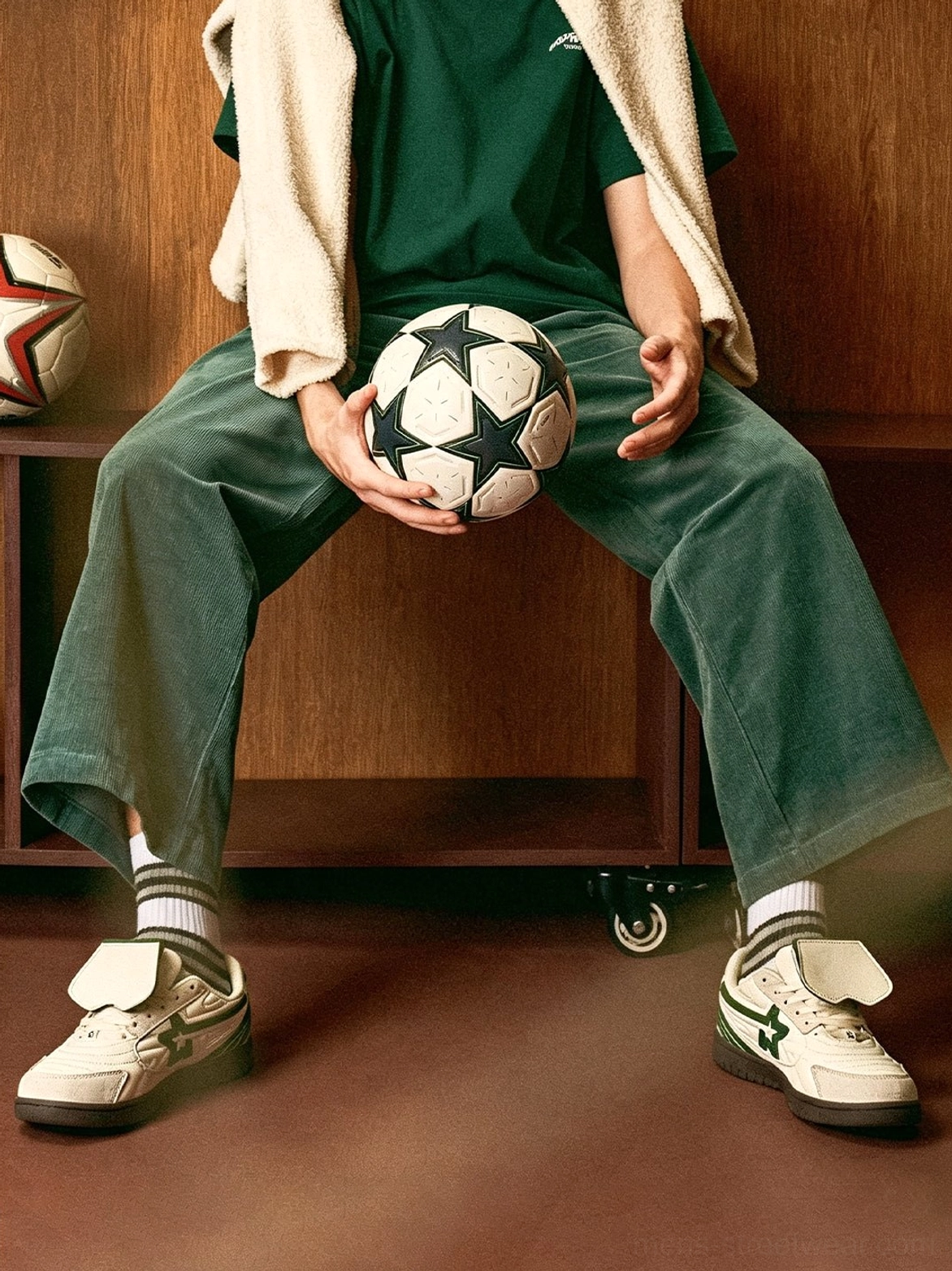 Male model sitting with a soccer ball while wearing white green low-top streetwear sneakers with flipped detachable tongues and striped socks