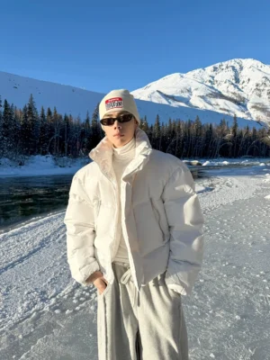 White high-neck stretch base layer styled under a white puffer jacket with grey wide-leg sweatpants in a snowy mountain riverside setting