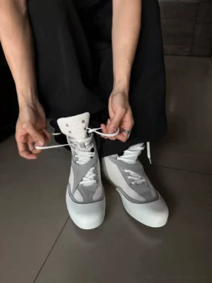 Model wearing white mesh high-top boxing boots with light gray overlays and loose black trousers while adjusting the front laces