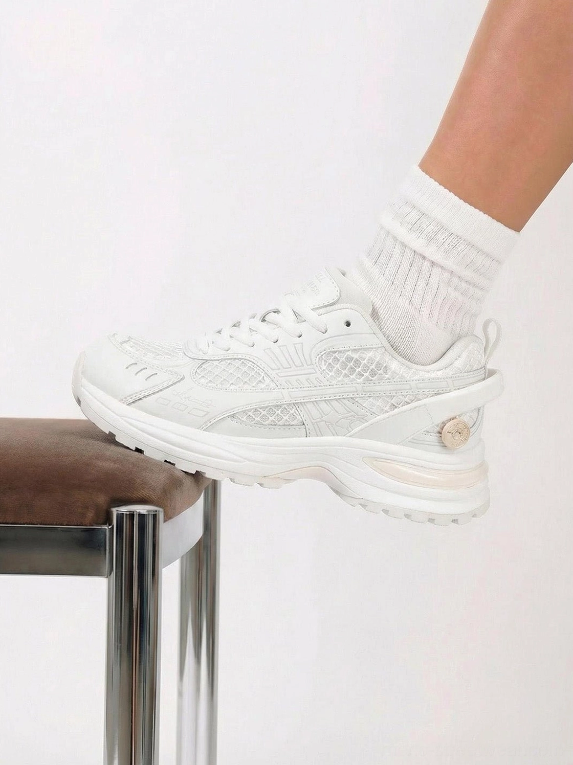 Single white mesh sneaker shown from a low side angle on a leather stool, highlighting the textured upper and thick sculpted sole