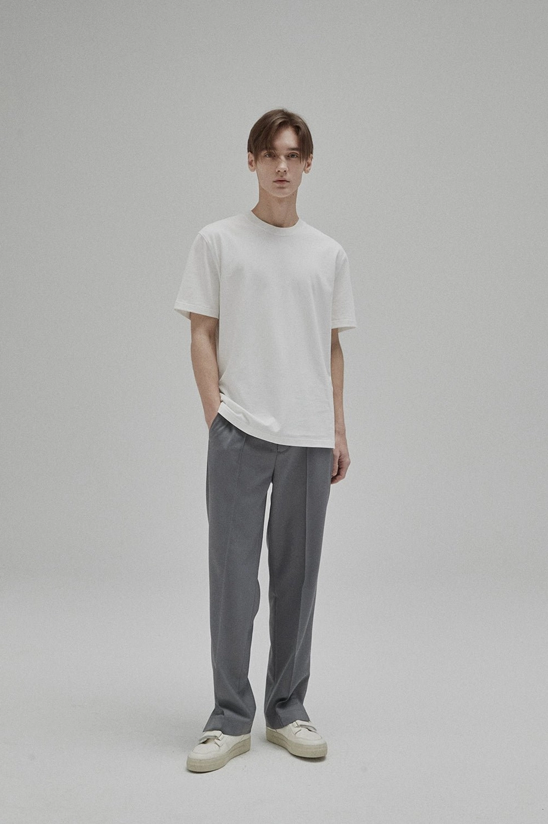 Full-body male model wearing a minimalist white short sleeve cotton T-shirt with grey tailored pants and casual sneakers