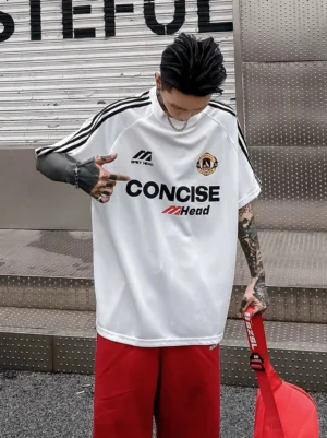 White oversized football jersey T-shirt with black triple shoulder stripes and collegiate front graphics worn in an industrial streetwear setting