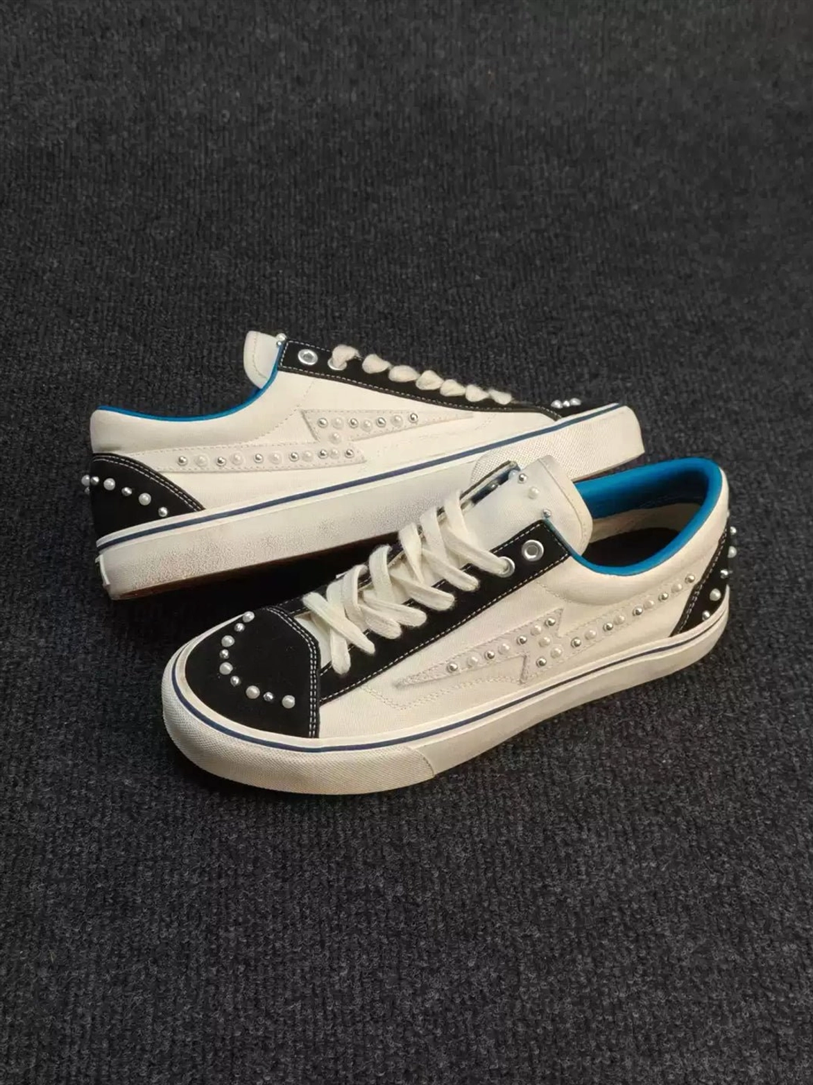 Side profile of white low-top pearl-studded skate sneakers featuring star-shaped stitching, black contrast panels, blue sole trim, and a rubber outsole