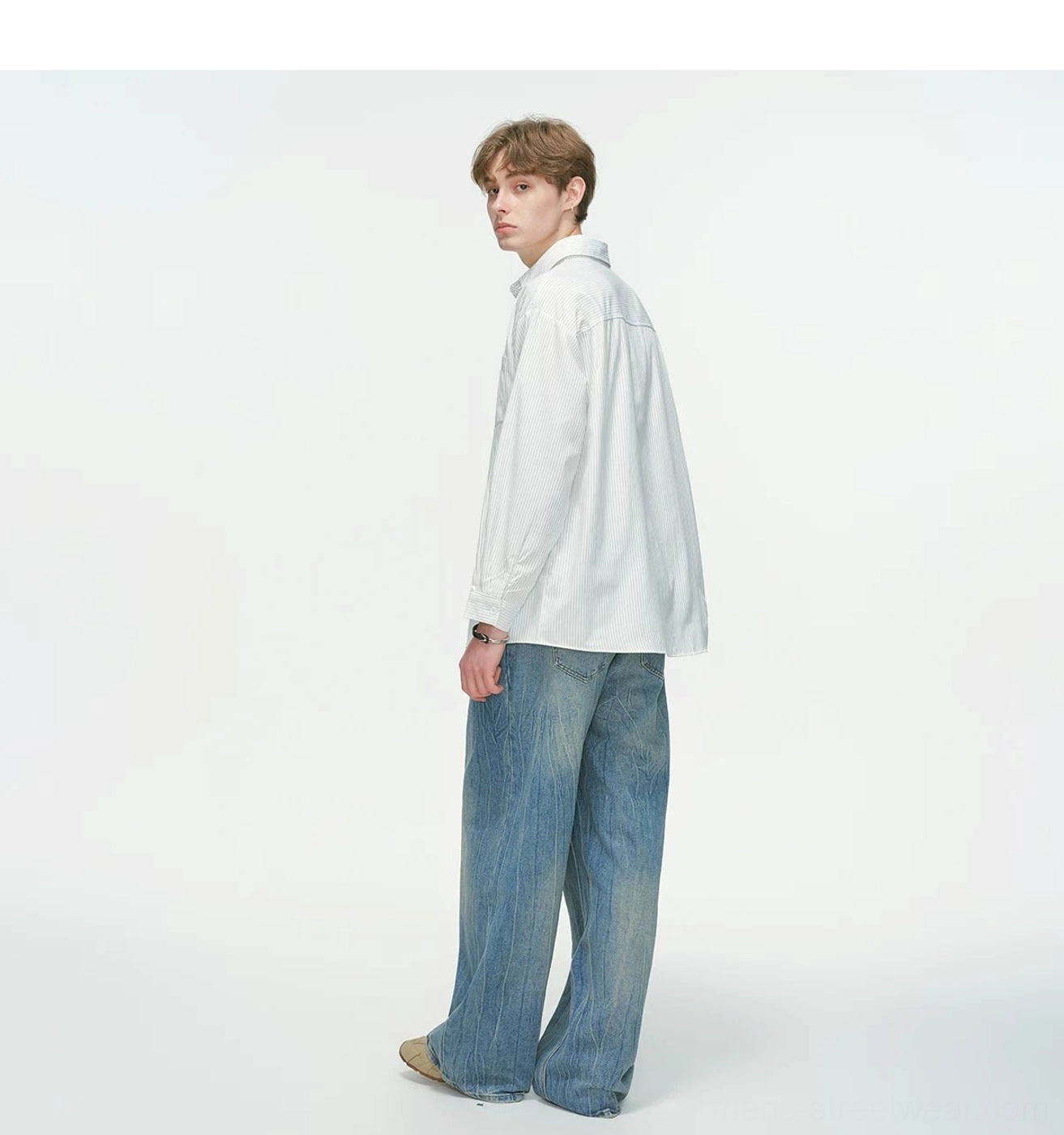 Side rear view of a male model wearing a loose white streetwear shirt with a clean back, relaxed hem, and wide-leg blue jeans