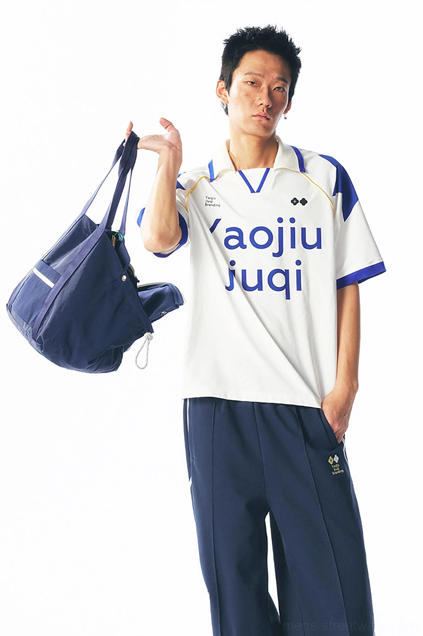 Front full-body view of a white retro printed football jersey with blue trim, navy sport pants, and a dark duffel bag