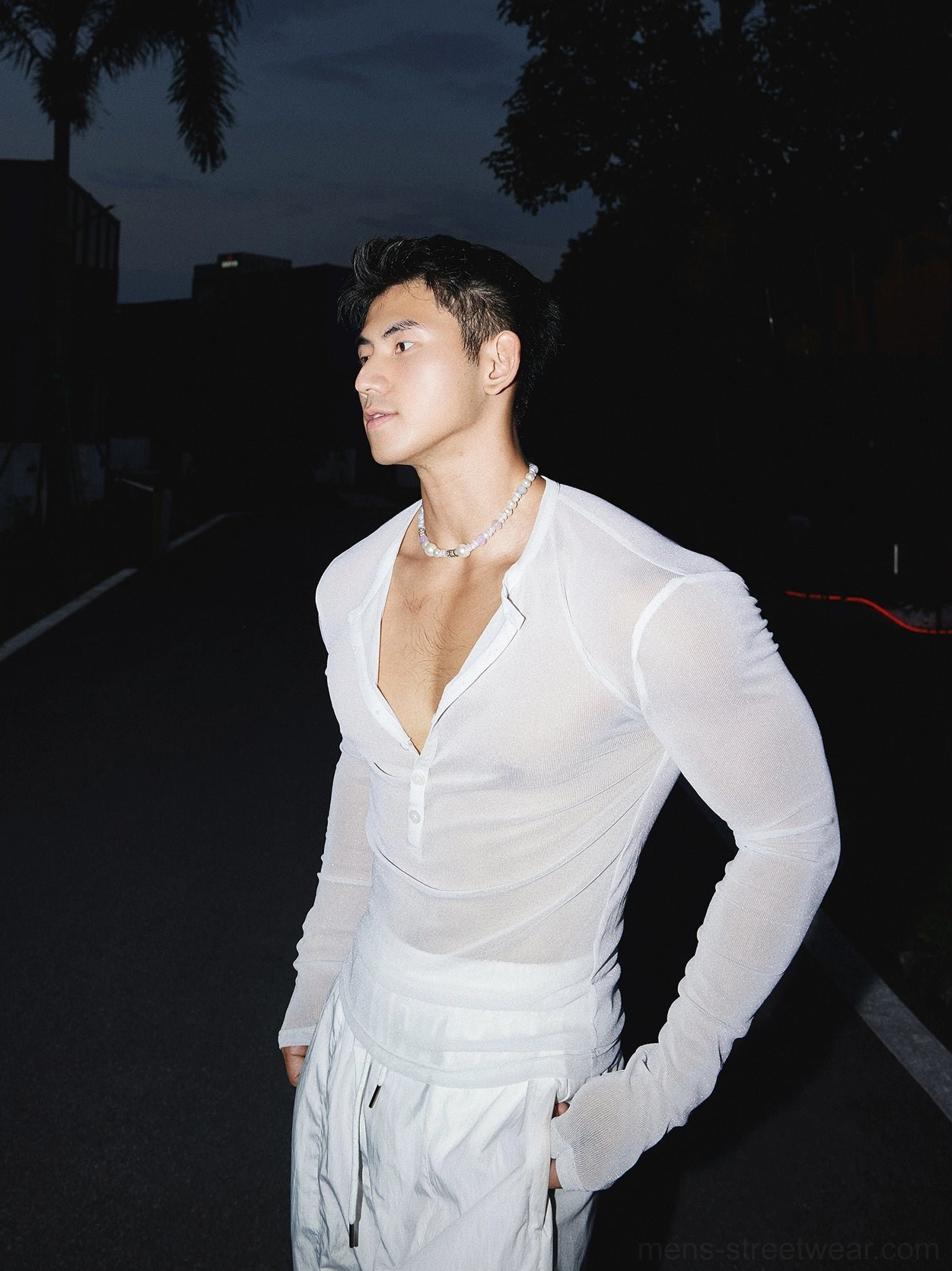Side full-length view of a male model wearing a white sheer sparkly Henley long sleeve top with wide-leg trousers